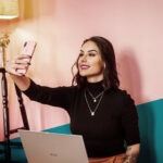 Junge Frau mit dunklen Haaren und Laptop auf dem Schoß macht ein Selfie vor einer pinken Wand