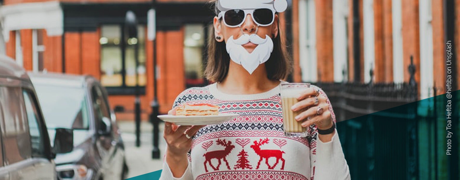 Frau mit Sonnenbrille, Weihnachtsmannmütze und weißem Kostüm-Bart, die einen Milchkaffe und ein Stück Kuchen in der Hand hält, steht auf der Straße