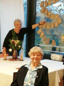 Zwei ältere Damen schauen in die Kamera. Eine von ihnen zeigt auf einen Adventskalender, der im Fenster hängt, die andere sitzt