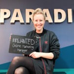 Junge blonde Frau sitzt vor einer Wand mit dem Schriftzug "Paradies" und hält eine Tafel, auf der steht "#wanted Online Marketing Manager"