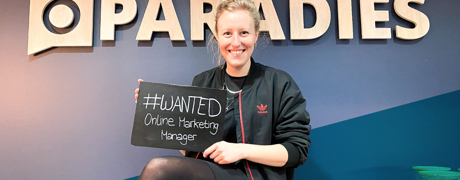 Junge blonde Frau sitzt vor einer Wand mit dem Schriftzug "Paradies" und hält eine Tafel, auf der steht "#wanted Online Marketing Manager"