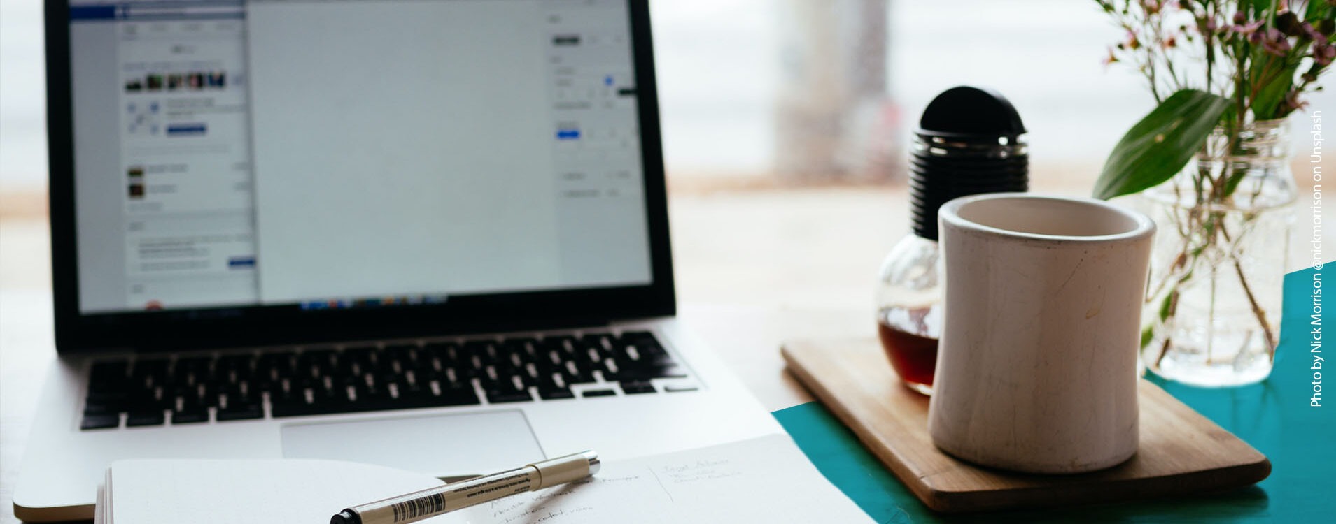 Schreibtisch mit einem Laptop, einem geöffneten Notizbuch mit Stift und einer Teetasse sowie Blumen